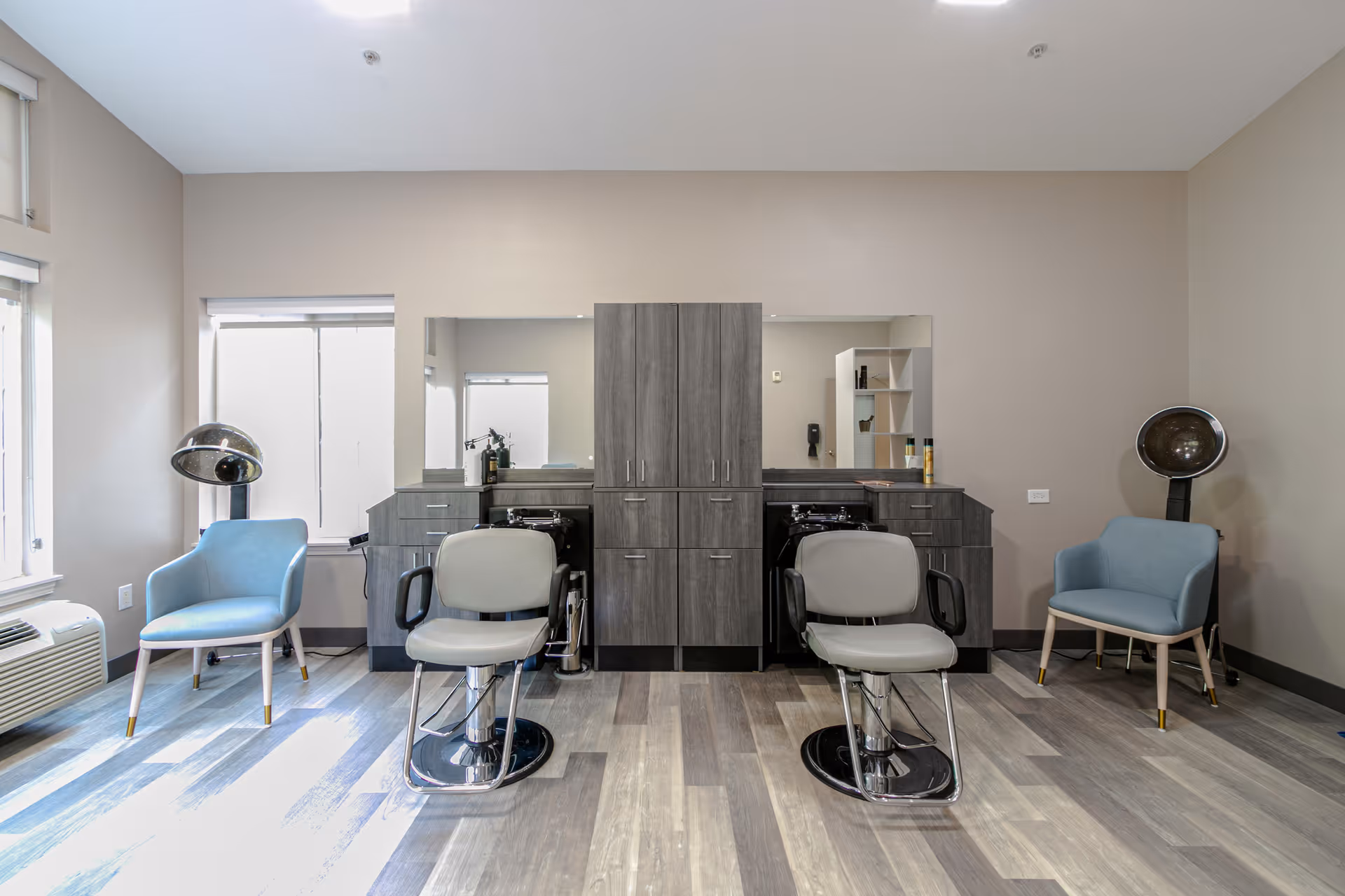 A modern hair salon area with two styling chairs in front of a large mirror and cabinetry. There are two blue armchairs on either side, each next to a vintage hair dryer. The room has light-colored walls and wood-patterned flooring, with windows allowing natural light to enter.