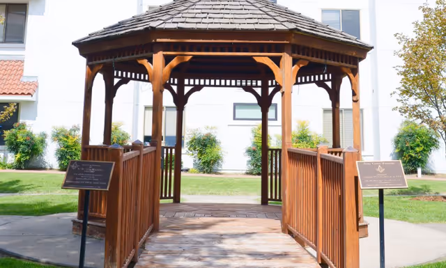 A wooden gazebo with a shingled roof and ramped walkway set in a grassy courtyard in front of a building.