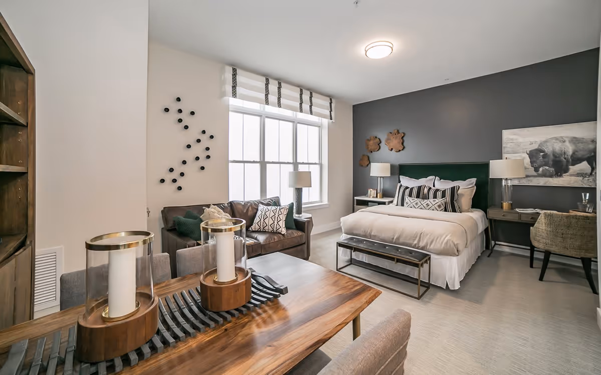 A modern and cozy studio apartment featuring a neatly made bed with decorative pillows against a dark accent wall, a small wooden desk with a chair, a brown leather couch with patterned cushions near a large window with white and gray striped valance, and a wooden dining table with two large candle holders in the foreground.