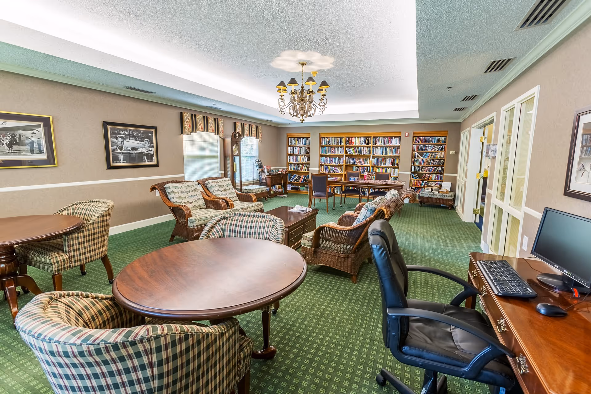 A cozy common area in a senior living facility featuring green patterned carpet, several round wooden tables with plaid upholstered chairs, wicker armchairs with cushions, a wooden coffee table, a computer desk with a black office chair, and a large bookshelf filled with books along the back wall. The room is softly lit by a chandelier and natural light from windows with valances. Framed black and white photographs hang on the beige walls.