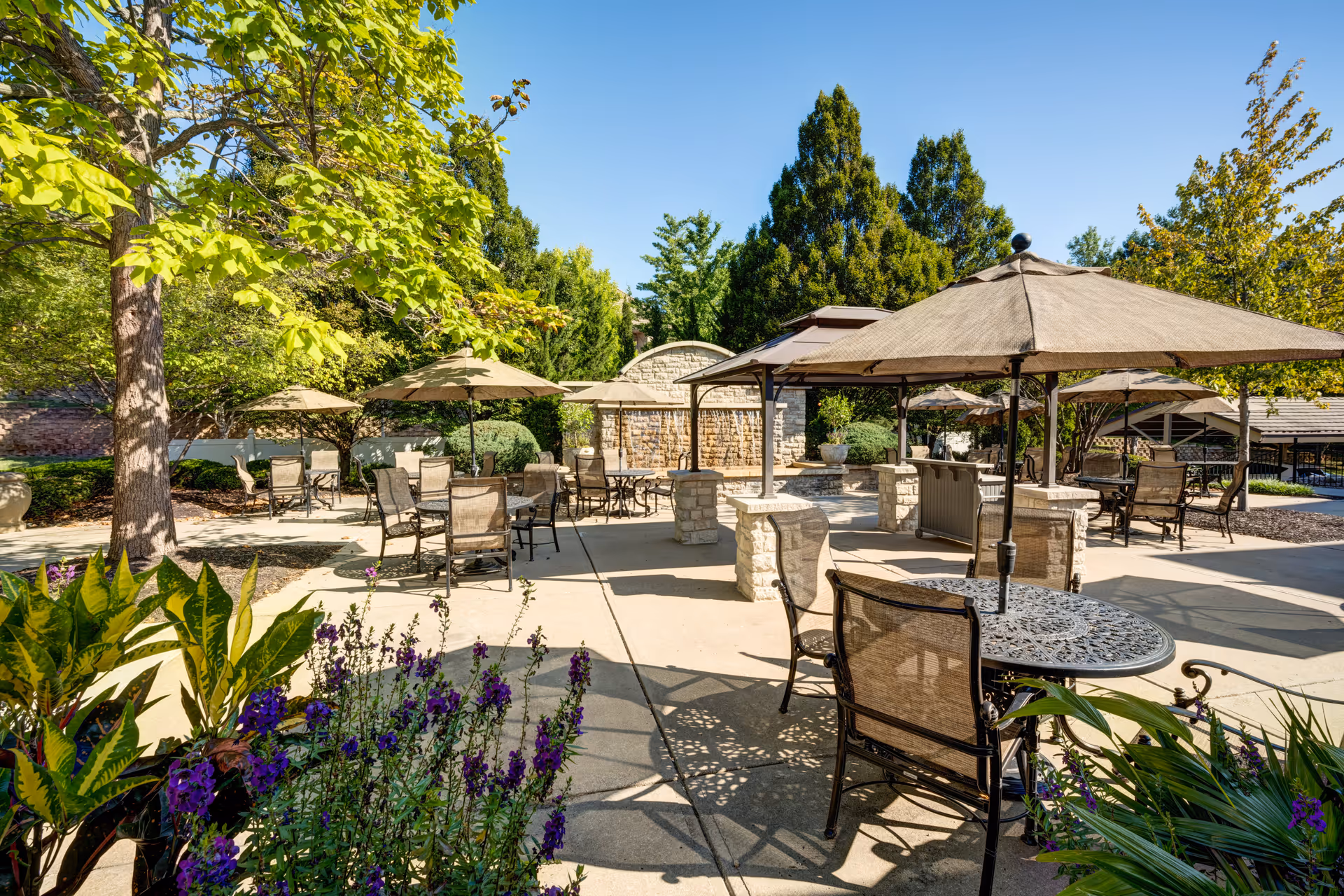 Outdoor patio area at Town Village of Leawood with multiple tables and chairs under large umbrellas, surrounded by trees, plants, and a stone water feature in the background under a clear blue sky.