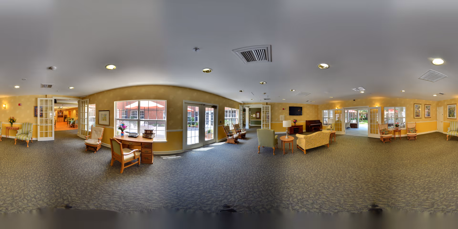 A spacious, well-lit common area in a senior living facility with multiple seating arrangements including armchairs, sofas, and small tables. The room features large windows and glass doors letting in natural light, beige walls with framed artwork, and carpeted flooring. There are also lamps, a television mounted on the wall, and floral decorations on tables. The area appears welcoming and comfortable for socializing or relaxing.