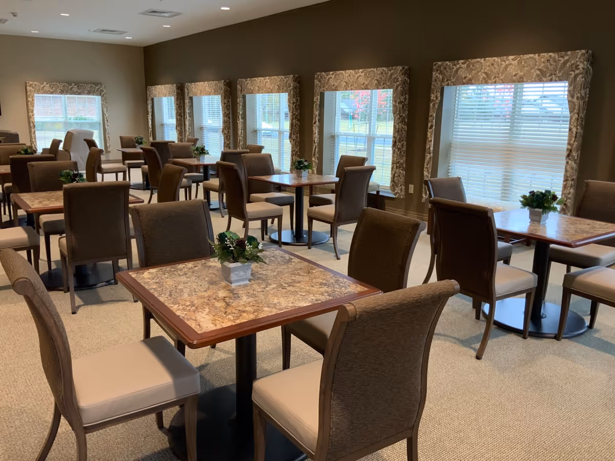 A dining room with multiple square tables, each surrounded by four cushioned chairs. The tables have a marble-like surface and small potted plants as centerpieces. The room has several large windows with patterned curtains, allowing natural light to brighten the space.
