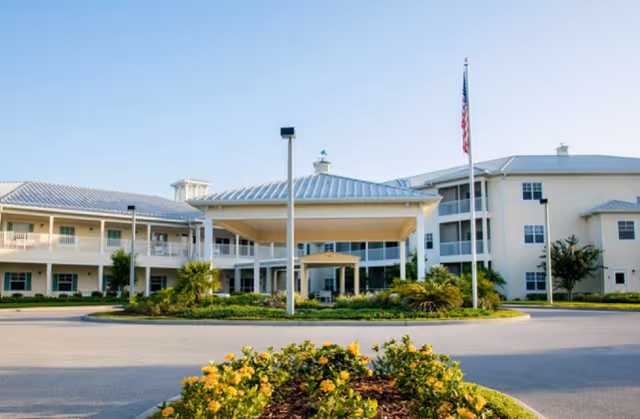 Front entrance of a multi-story senior living building with a covered porte-cochère, circular driveway, landscaping, and an American flag.