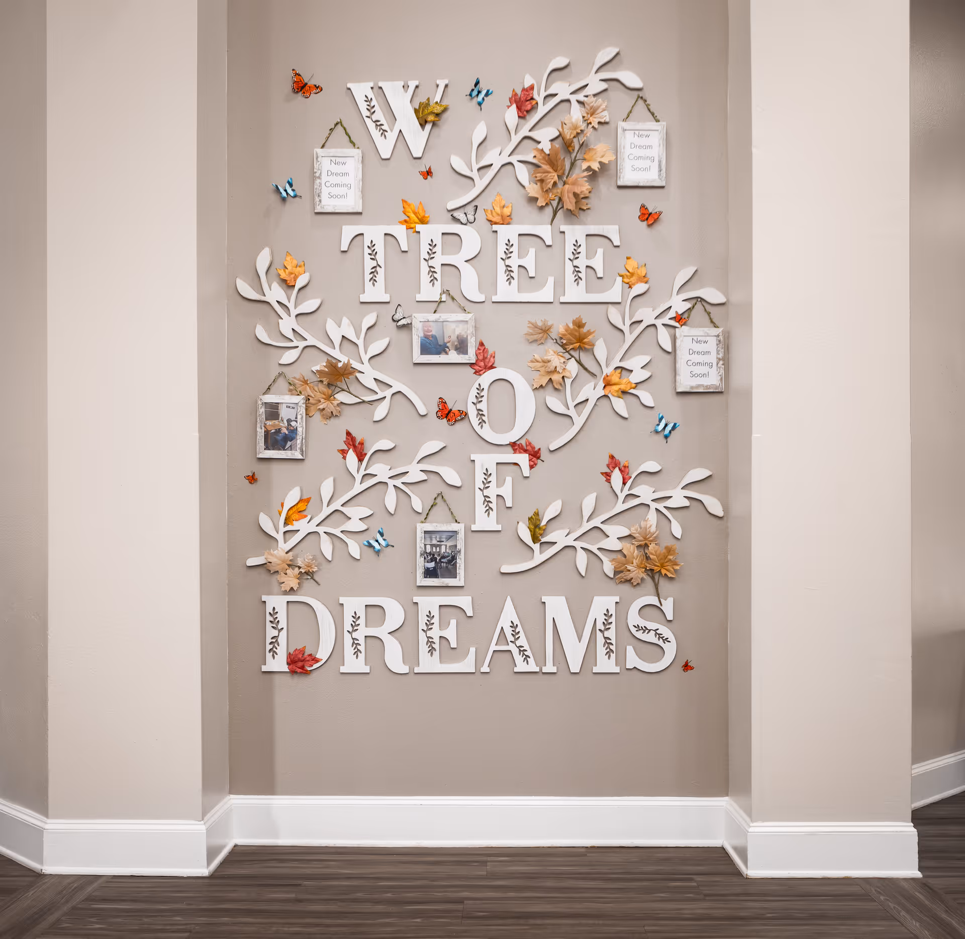 Wall decoration with large white letters spelling 'W TREE OF DREAMS' surrounded by white leafy branches, autumn leaves, colorful butterflies, and small framed pictures and signs that say 'New Dream Coming Soon!'