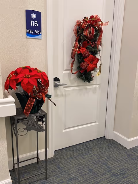 A white door decorated with a festive holiday wreath featuring red ribbons and greenery. To the left of the door is a small metal stand holding a pot of red poinsettia flowers. A blue room number sign above the door handle reads '116 May Bice'. The surrounding walls are light-colored and the floor is carpeted in a blue pattern.