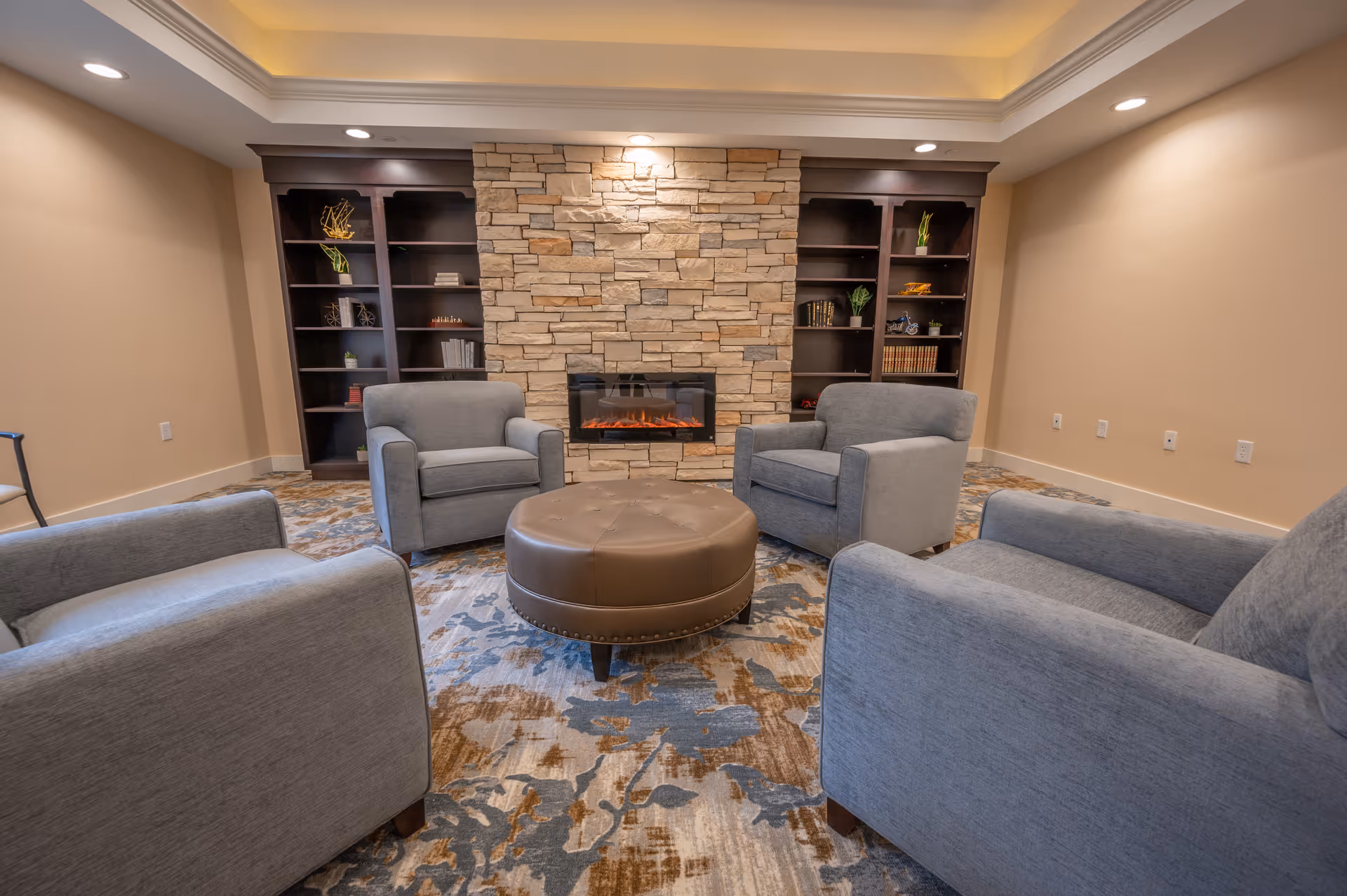 A cozy living room with four gray armchairs arranged around a round brown leather ottoman. The room features a stone fireplace with a modern electric fire and built-in dark wood bookshelves on either side, decorated with books and small plants. The ceiling has recessed lighting and a soft cove light, and the floor is covered with a patterned carpet in shades of blue, brown, and beige.
