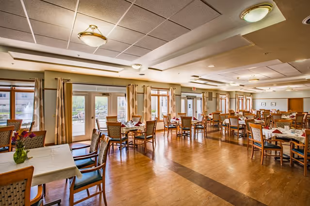 A spacious dining room with multiple wooden tables and chairs arranged neatly. Each table is set with white tablecloths, napkins, and some have small flower vases. Large windows and glass doors along one wall allow natural light to fill the room, with curtains framing the windows. The ceiling has recessed lighting and decorative light fixtures, and the floor is wooden.