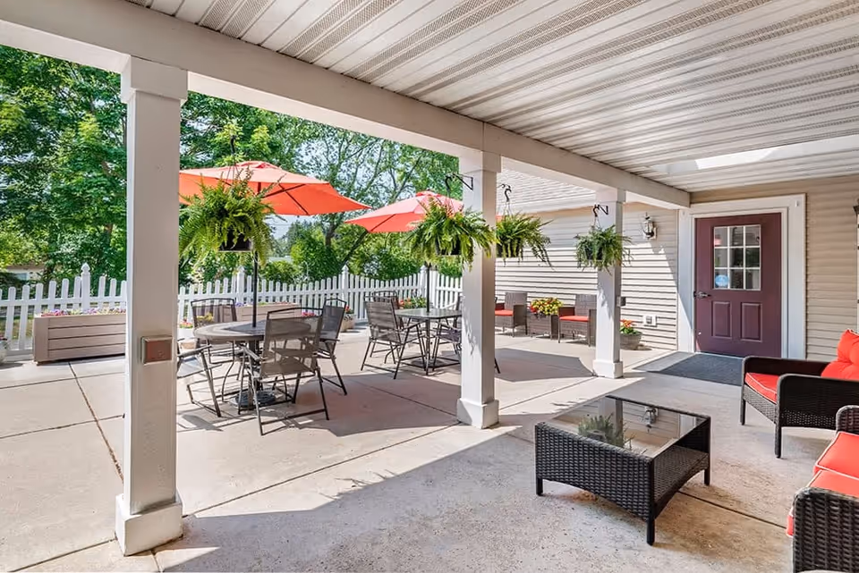 Covered outdoor patio area with hanging ferns, several tables with chairs and red umbrellas, and cushioned wicker seating with a glass-top coffee table. A white picket fence and green trees are visible in the background.