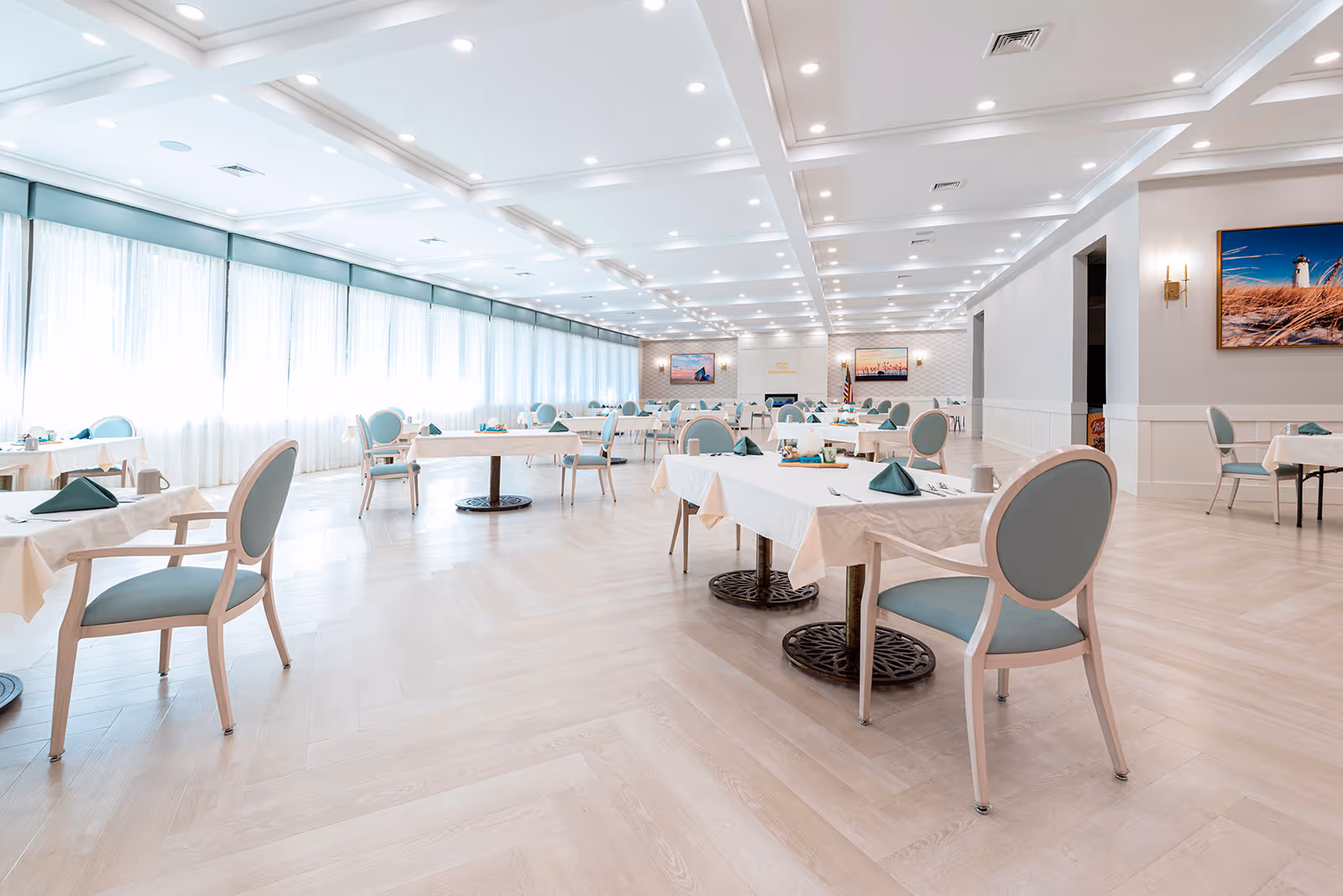 Bright, spacious dining room with neatly set white-clothed tables and teal-upholstered chairs.