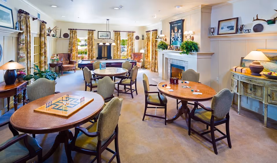 A bright and cozy common room with multiple round wooden tables and cushioned chairs arranged for games and social activities. The room features a fireplace with plants and artwork above it, large windows with patterned curtains letting in natural light, and comfortable armchairs near the windows. The walls are decorated with framed pictures and shelves with decorative items.