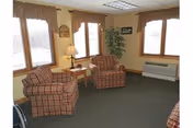 Cozy sitting room with two red plaid armchairs, a side table with a lamp, a potted plant, and three windows.