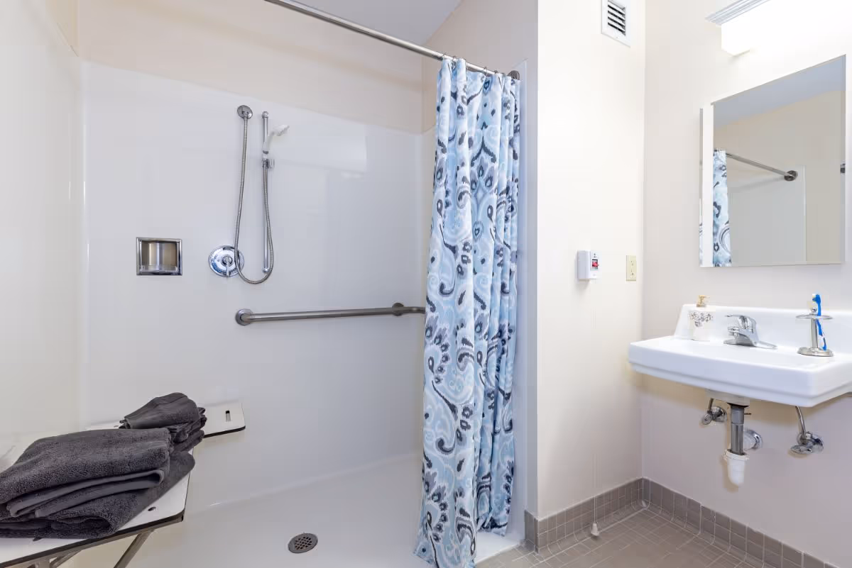 A clean bathroom with a walk-in shower featuring a handheld showerhead and a blue patterned shower curtain. There is a foldable shower seat with folded dark gray towels on it. A white sink with a mirror above it is visible on the right side, along with a soap dispenser and a toothbrush holder.