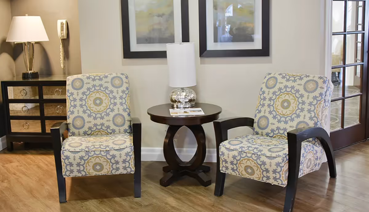 Two patterned armchairs with wooden arms and legs are positioned on either side of a round wooden side table with a decorative lamp on top. Behind the chairs, there are two framed abstract paintings on a light-colored wall. To the left, there is a dark wooden chest of drawers with a lamp and a wall-mounted telephone. To the right, a glass-paneled door leads to another room.