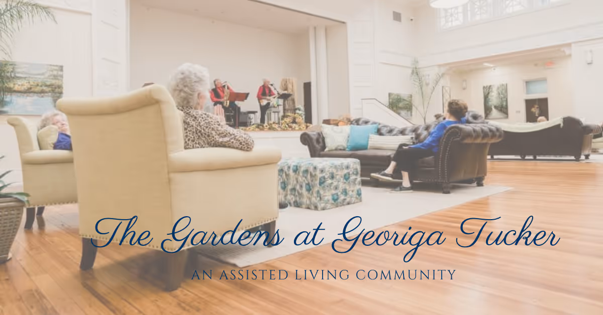A spacious living room area in an assisted living community with elderly residents seated on armchairs and sofas, watching a live musical performance on a small stage. The room has wooden floors, large windows, and light-colored walls with artwork.