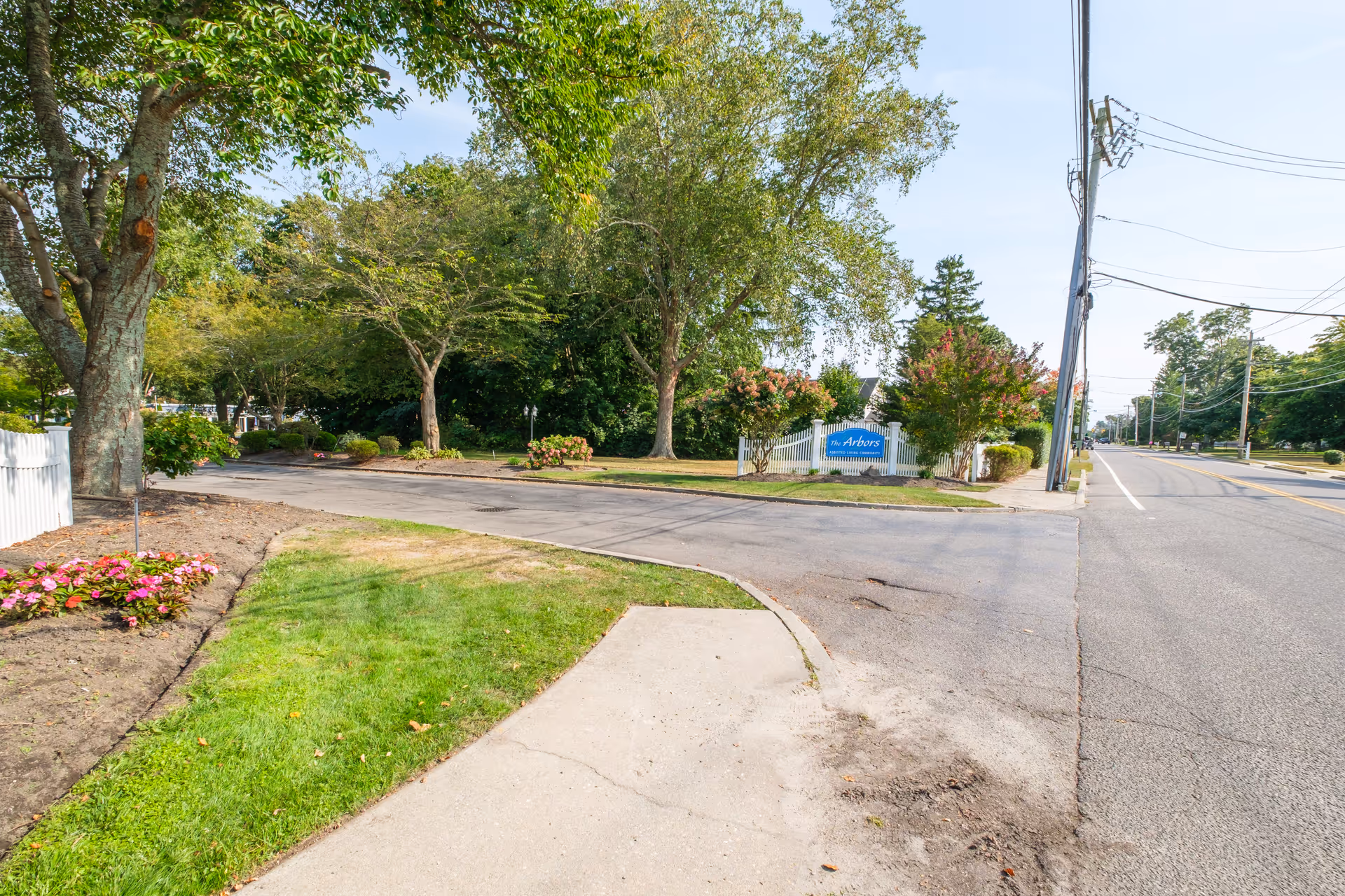 View of the entrance to The Arbors assisted living community featuring a paved driveway, green grass, flower beds, trees, and a white picket fence with a blue sign displaying the facility name.