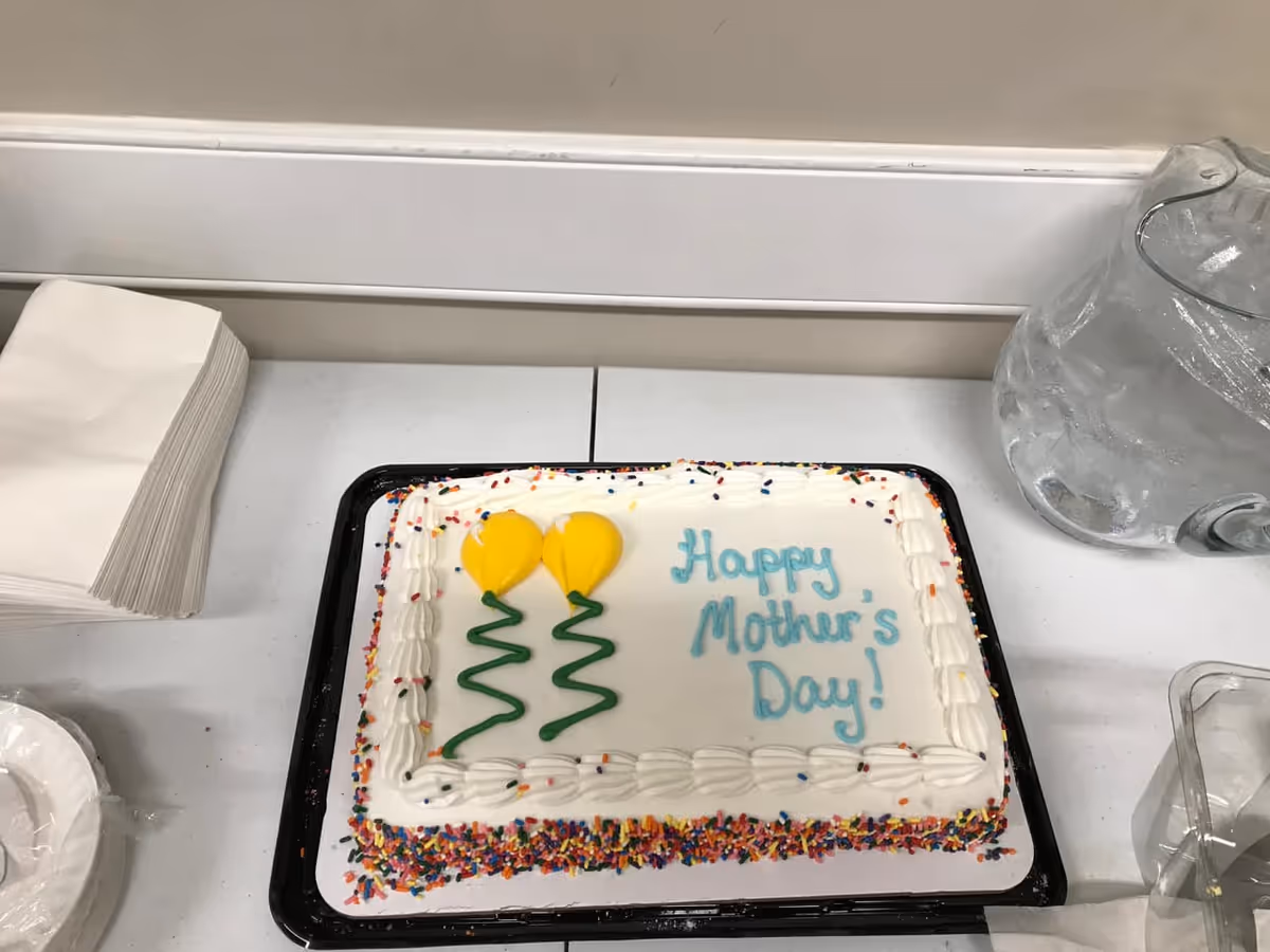 A rectangular cake with white frosting decorated with colorful sprinkles around the edges and two yellow balloon designs. The cake has the message 'Happy Mother's Day!' written in blue icing. The cake is placed on a white table next to a stack of white napkins and a clear pitcher of water with ice.