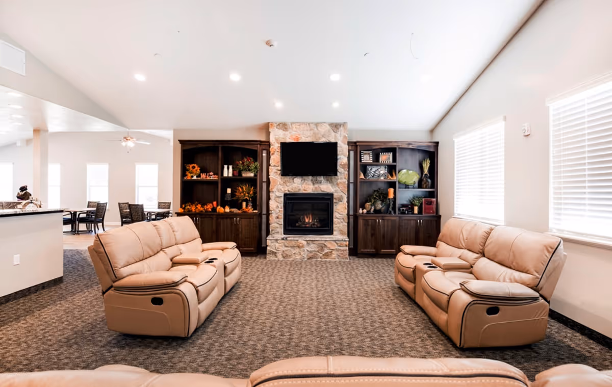 A bright and spacious living room with two beige leather reclining sofas facing each other. In the center of the far wall is a stone fireplace with a mounted flat-screen TV above it. On either side of the fireplace are dark wooden built-in shelves decorated with plants, books, and seasonal decor. Large windows with white blinds allow natural light to fill the room. The carpet is patterned in neutral tones, and the ceiling has recessed lighting.