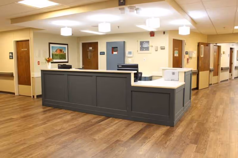 Reception desk area inside a senior living facility with wood flooring, beige walls, and multiple doors in the background. The desk is gray with a white countertop, and there are computer monitors and a small printer on it. Ceiling lights illuminate the space.