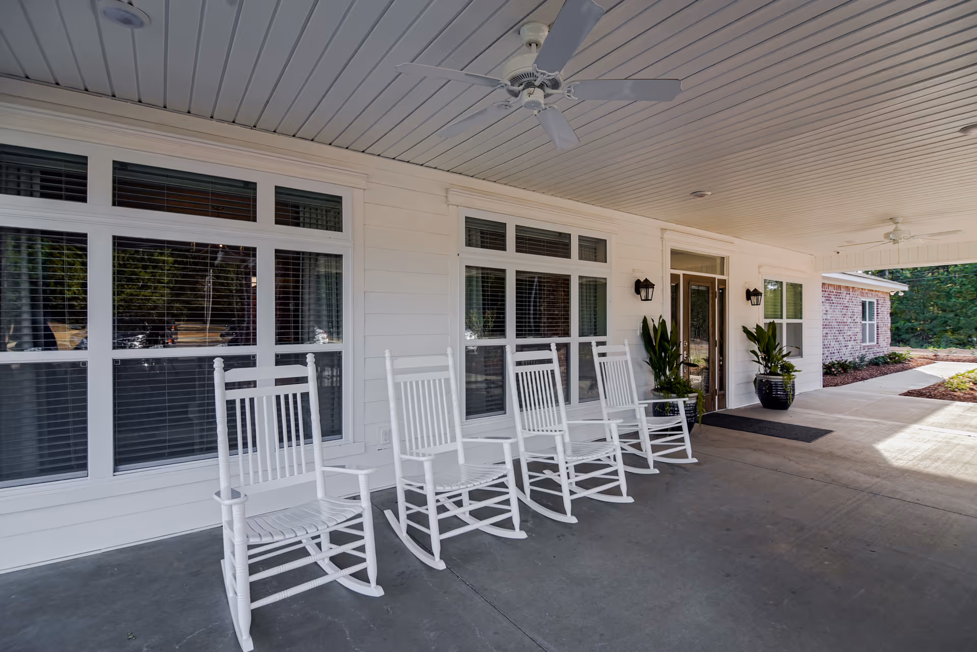 Covered porch area with five white rocking chairs lined up against the white exterior wall of a building. The porch has a ceiling with two ceiling fans and two black wall-mounted lantern lights near a glass door entrance. There are two large potted plants on either side of the door and windows with blinds behind the chairs. The ground is concrete and the area is well-lit with natural light.