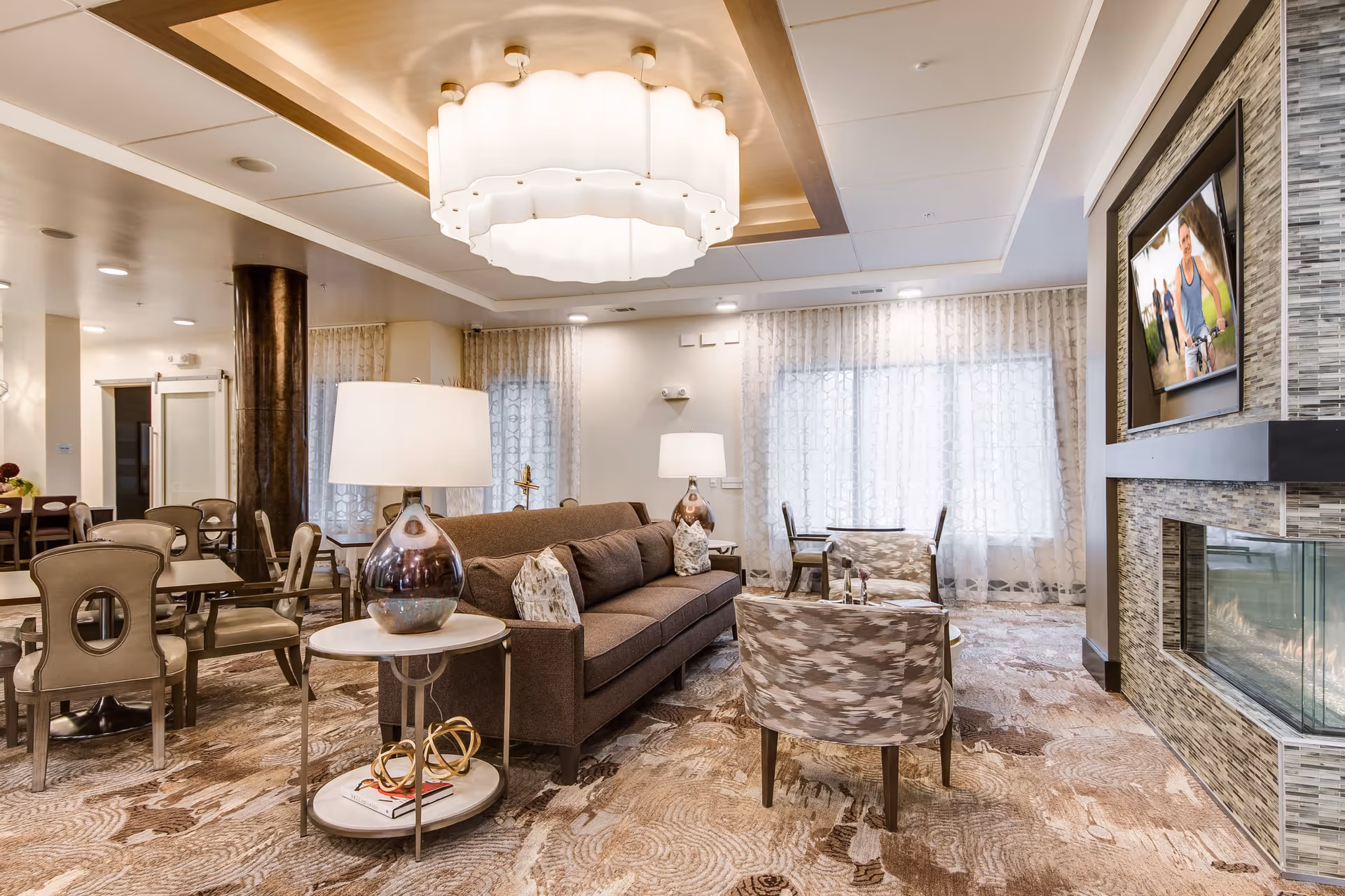 A cozy senior living facility common area with a brown sofa, patterned armchairs, side tables with lamps, a modern fireplace, and a wall-mounted TV. The room has large windows with sheer curtains, a decorative ceiling light fixture, and a patterned carpet.