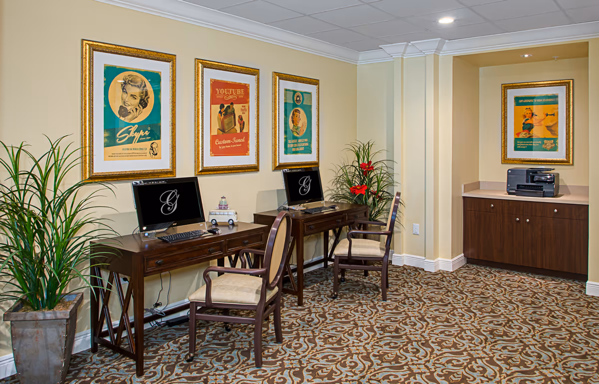 A cozy computer corner in a senior living facility with two wooden desks, each holding a computer and a chair. The walls are decorated with four vintage-style framed posters. There are two potted plants on either side of the desks, and a printer sits on a built-in cabinet in a recessed wall area. The room has a patterned carpet and soft yellow walls.