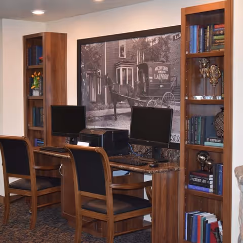 A small computer workstation area with two desktop computers and black chairs. The desks and bookshelves are made of wood, and the bookshelves contain books and decorative items. A large black and white historical photograph of a horse-drawn carriage and buildings is mounted on the wall behind the computers.