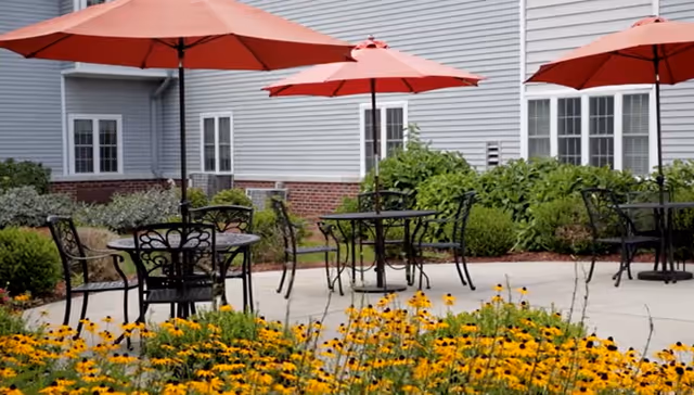 Outdoor patio area with black metal tables and chairs under red umbrellas, surrounded by yellow flowers and green shrubs in front of a light gray building with white-framed windows.