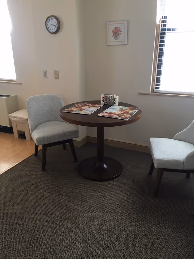 Small dining nook with a round wooden table set with placemats between two upholstered chairs by windows.