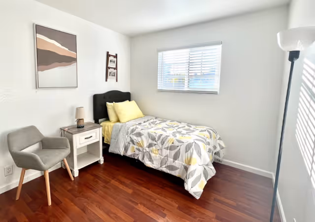 A bright, clean bedroom with a single bed featuring a floral patterned bedspread in shades of gray and yellow. The bed has a black tufted headboard and yellow pillows. Next to the bed is a white nightstand with a small lamp, and a gray upholstered chair is positioned nearby. A window with white blinds lets in natural light, and a floor lamp stands in the corner. The room has wooden flooring and white walls with minimal decor, including a framed abstract artwork and a small hanging photo display.