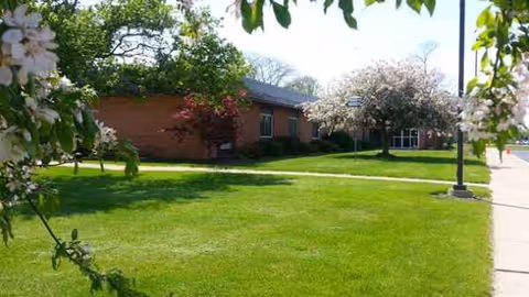 Lawn, brick building, trees, and blooming shrubs frame Urbana Health and Rehab Center outdoor area.