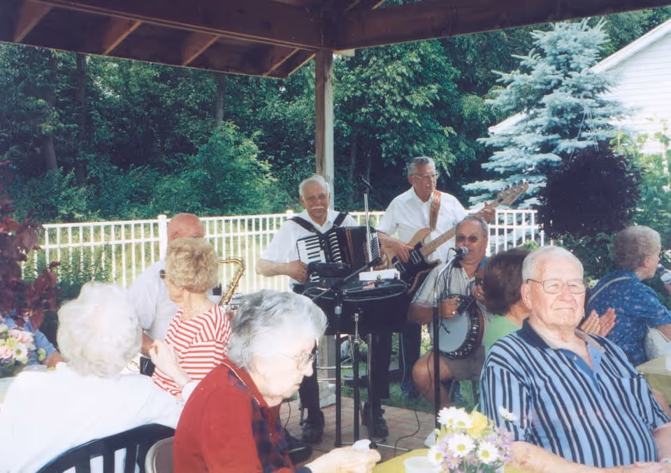 A group of elderly people seated outdoors under a covered patio, enjoying a live musical performance by three musicians playing an accordion, guitar, and banjo. The setting is surrounded by greenery and a white fence.