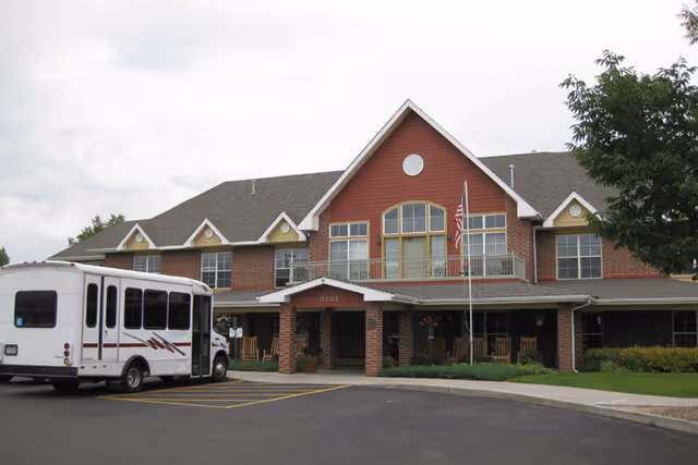 Exterior view of a two-story assisted living facility building with a covered entrance, several windows, an American flag on a flagpole, and a white shuttle bus parked in front.