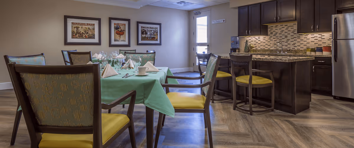 A dining area with a table covered in a green tablecloth set with napkins, glasses, and cups. Surrounding the table are wooden chairs with yellow cushions. In the background, there is a kitchen area with dark wood cabinets, a granite countertop island with stools, a stainless steel refrigerator, and a tiled backsplash. Three framed pictures hang on the wall, and a door with a window is visible.
