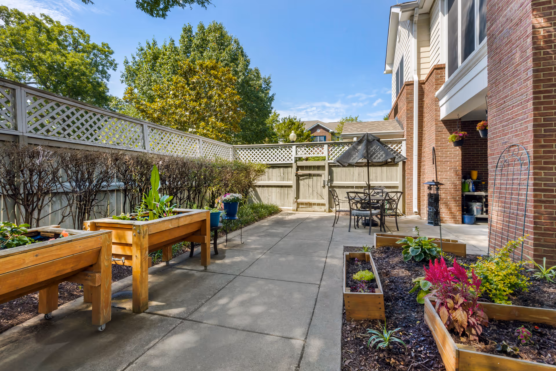 Sunny fenced courtyard with raised wooden planter boxes, a patio table and umbrella beside a brick building.