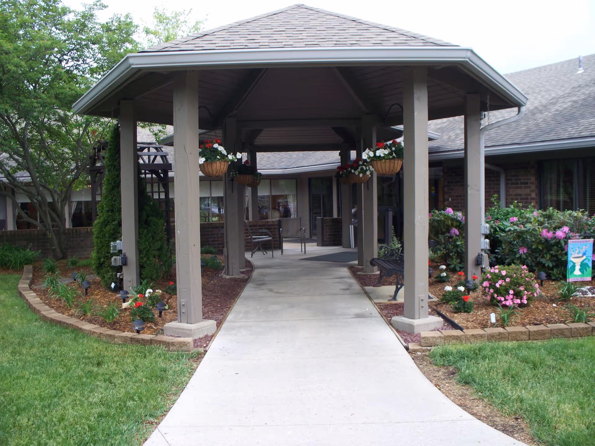 Covered walkway entrance to a building with hanging flower baskets and landscaped flower beds on either side. The building has brick walls and large windows, with benches placed along the walkway.