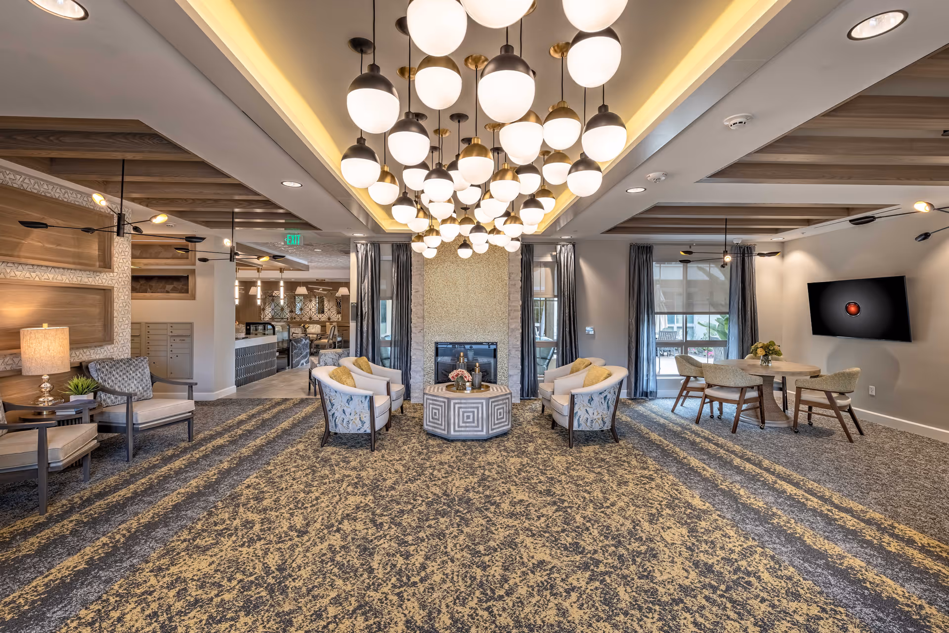 Spacious, well-lit senior living lounge with seating arranged around a central fireplace, chandeliers overhead, and a dining table to the right.