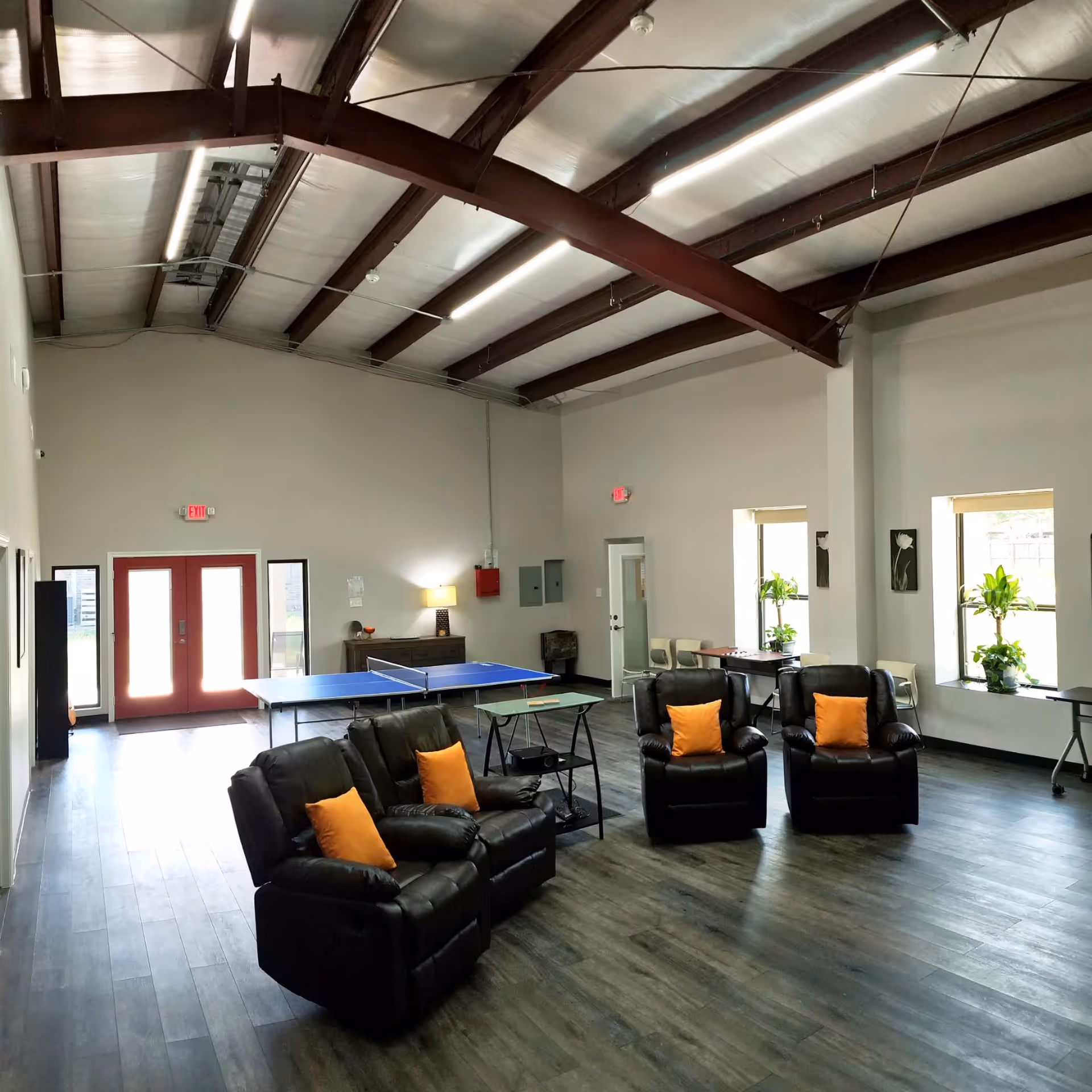 Spacious communal lounge with leather recliners accented by orange pillows, a ping-pong table, and a high exposed-beam ceiling.
