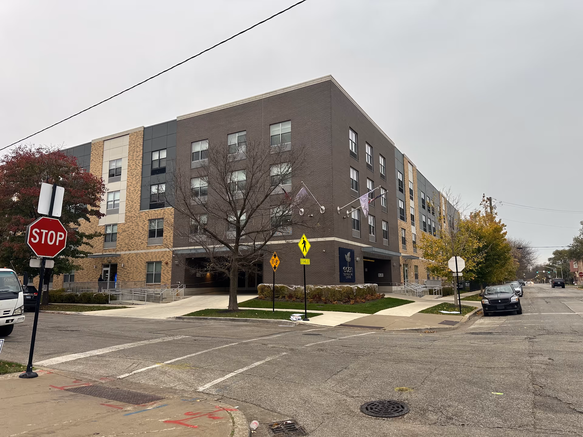 Exterior view of a modern four-story building at a street corner with a stop sign and pedestrian crossing signs. The building has a mix of dark and light brick facades with multiple windows. There is a sign on the building that reads 'eden supportive living'. Trees with autumn foliage are visible along the sidewalk.