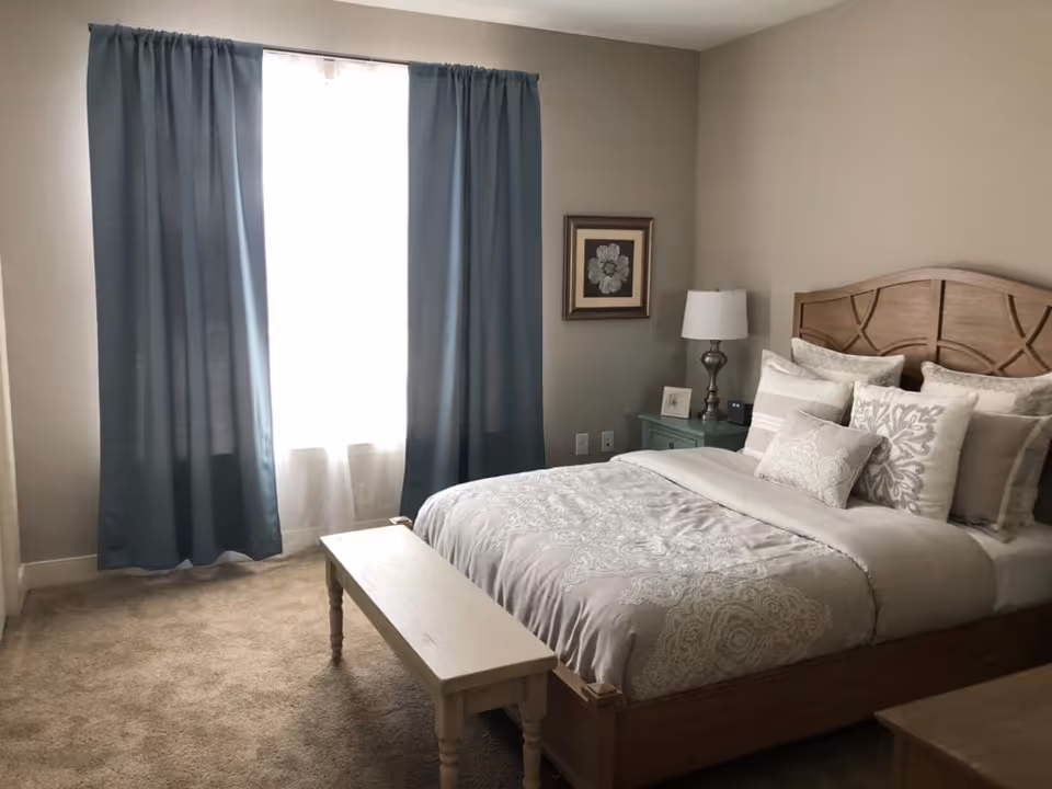 A cozy bedroom with a wooden bed frame and a neatly made bed featuring multiple decorative pillows and a patterned comforter. There is a wooden bench at the foot of the bed, a nightstand with a lamp, a framed flower artwork on the wall, and a window with blue curtains allowing natural light to enter the room.