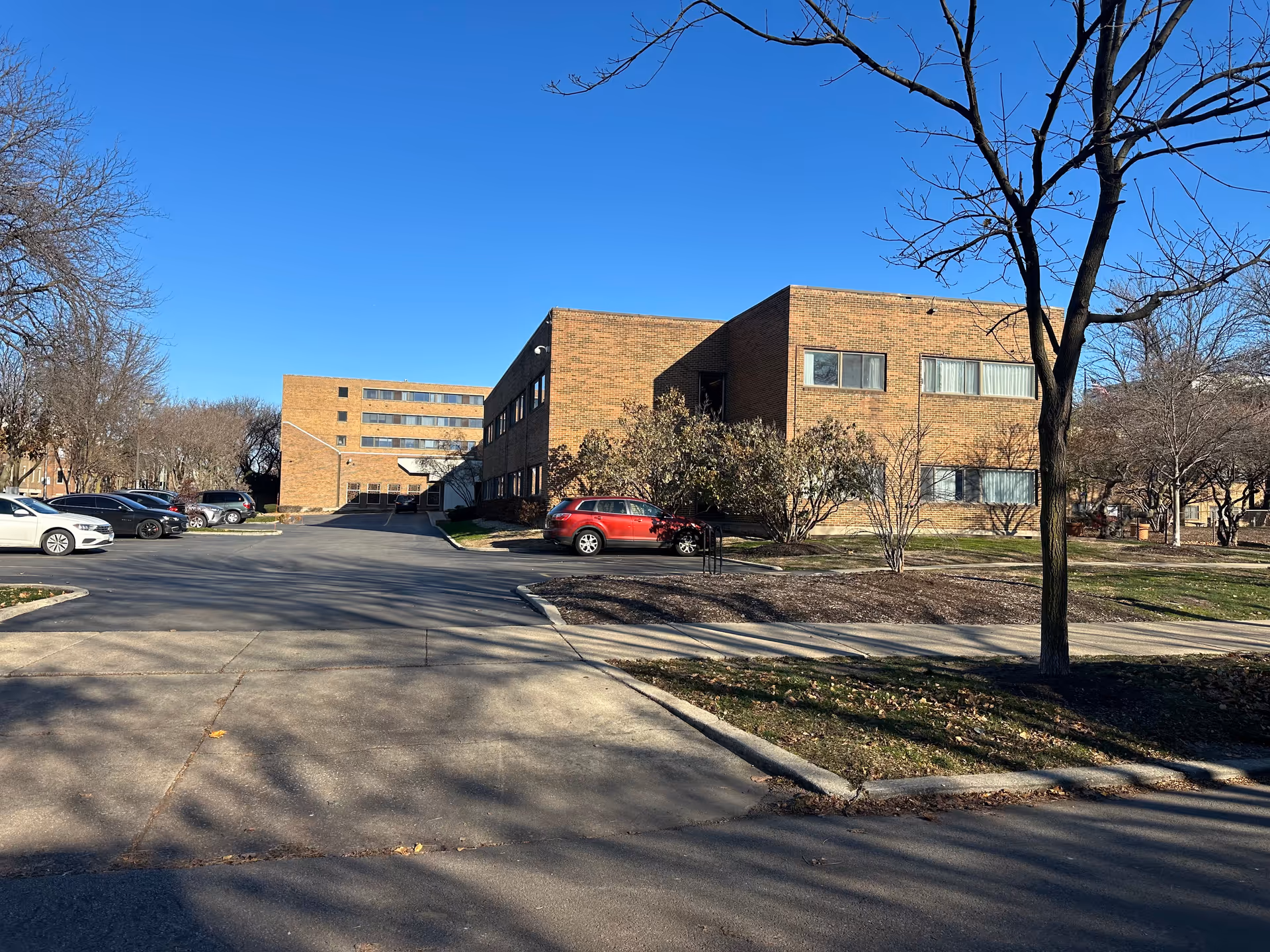 Exterior view of a low-rise brick senior living building with a parking lot, leafless trees, and a clear blue sky.