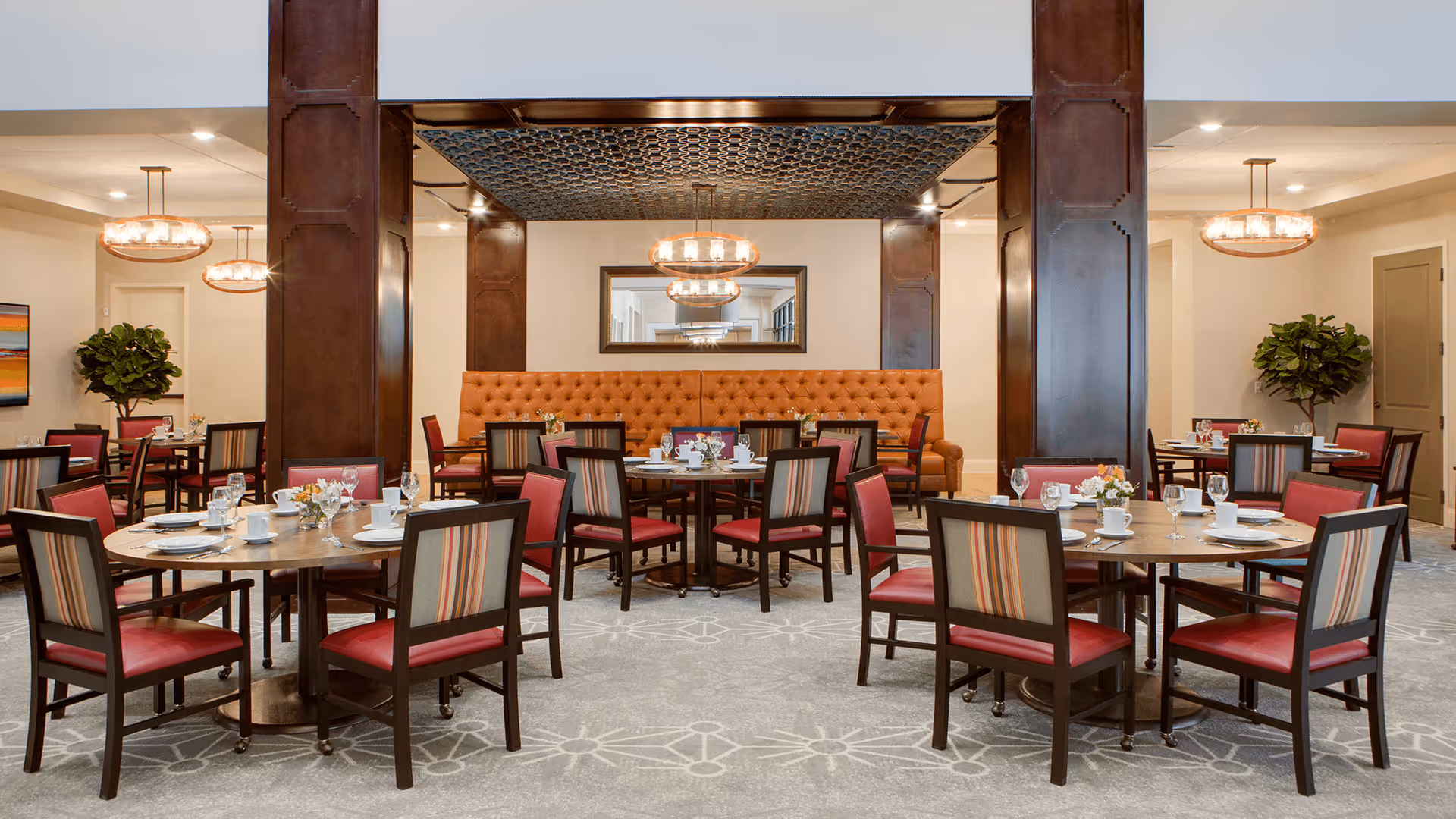 A well-lit dining room with round wooden tables set with white plates, cups, and glassware. The chairs have dark wooden frames with red seats and striped backs. There is a long orange tufted bench along the back wall, flanked by dark wooden columns. Modern chandeliers hang from the ceiling, and there are green potted plants in the corners of the room.