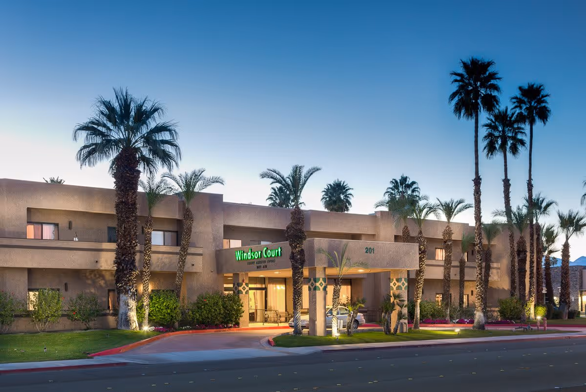 Exterior view of Windsor Court Assisted Living facility at dusk, featuring a two-story building with palm trees and well-maintained landscaping along the entrance driveway.