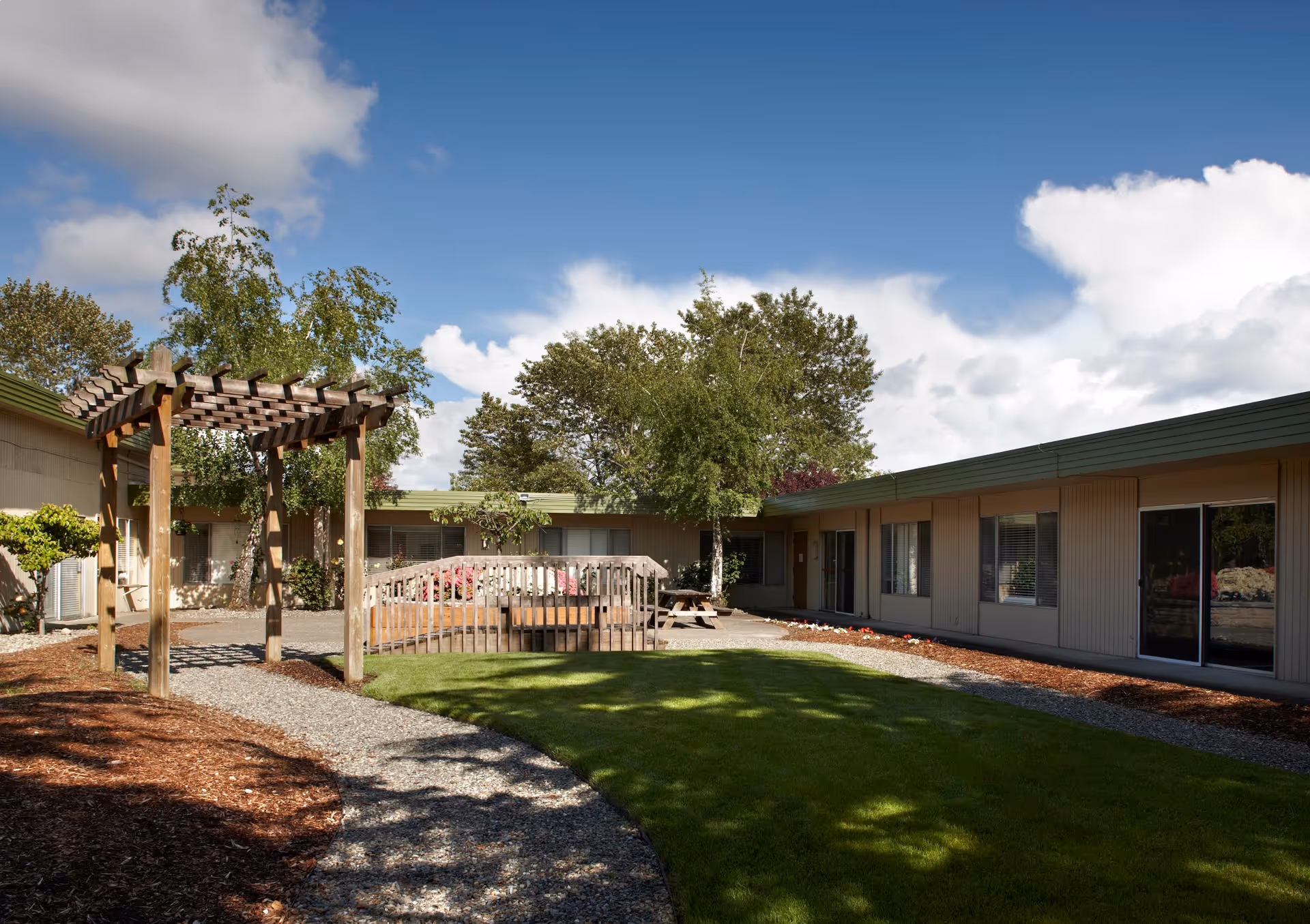 Outdoor courtyard area of a senior living facility with a wooden pergola, a small wooden bridge over a garden bed, picnic tables, green grass, trees, and a building with multiple windows surrounding the courtyard under a partly cloudy blue sky.