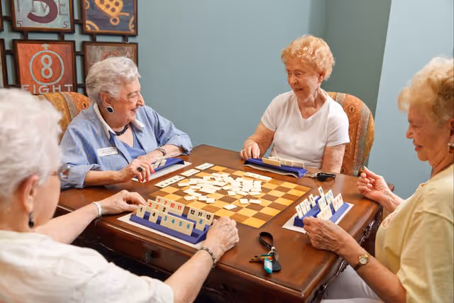 Four elderly women sitting around a wooden table playing a tile-based board game in a cozy room with light blue walls and framed artwork on the wall.