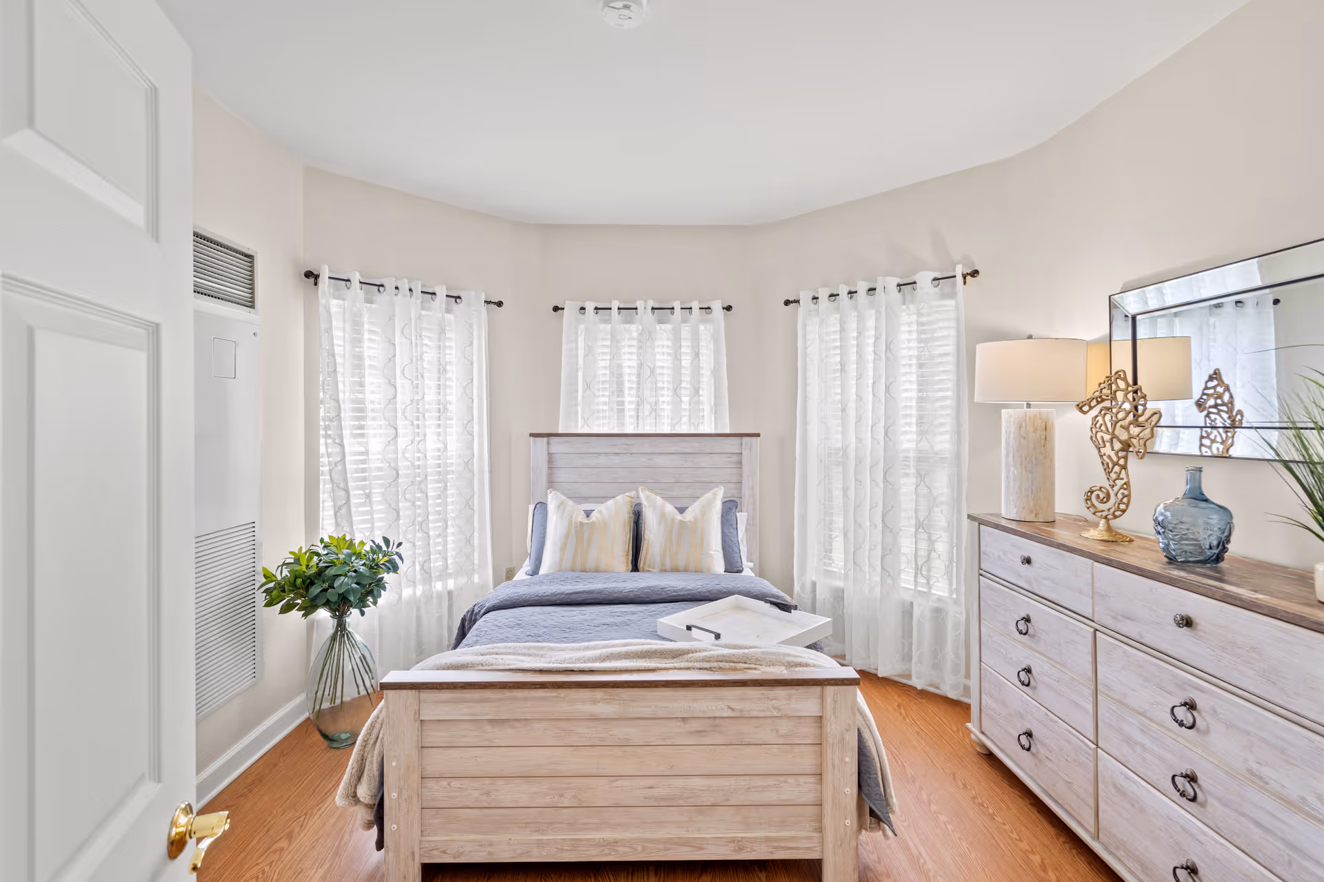 A bright bedroom with a wooden bed frame, a neatly made bed with blue and beige pillows, and a light blanket. The room has three windows with sheer white curtains, a wooden dresser with a lamp, decorative seahorse, blue vase, and a mirror above it. There is a glass vase with green plants on the floor near the windows.