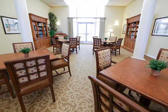 Bright communal dining/activity room with wooden tables and chairs, bookshelves, patterned carpet, and large windows.