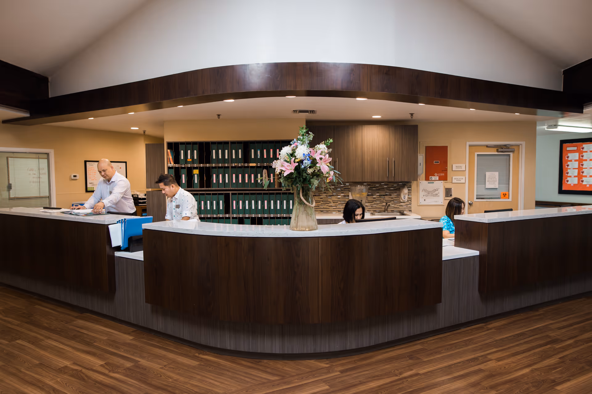Reception desk with staff working behind a curved wood counter and a vase of flowers in a senior care facility lobby.
