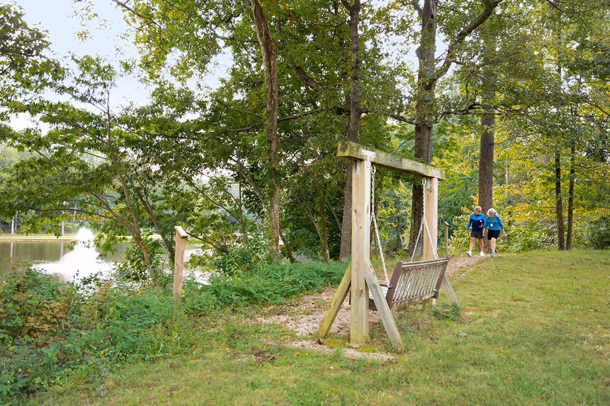 A peaceful outdoor scene at Aldersgate featuring a wooden swing bench on a grassy area near a walking path. Two elderly women are walking together along the path surrounded by trees and greenery. In the background, there is a pond with a water fountain and a small bridge partially visible through the trees.