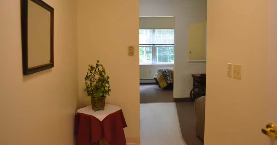 View down an interior hallway with a small table and potted plant, leading into a bedroom with a bed by a window.